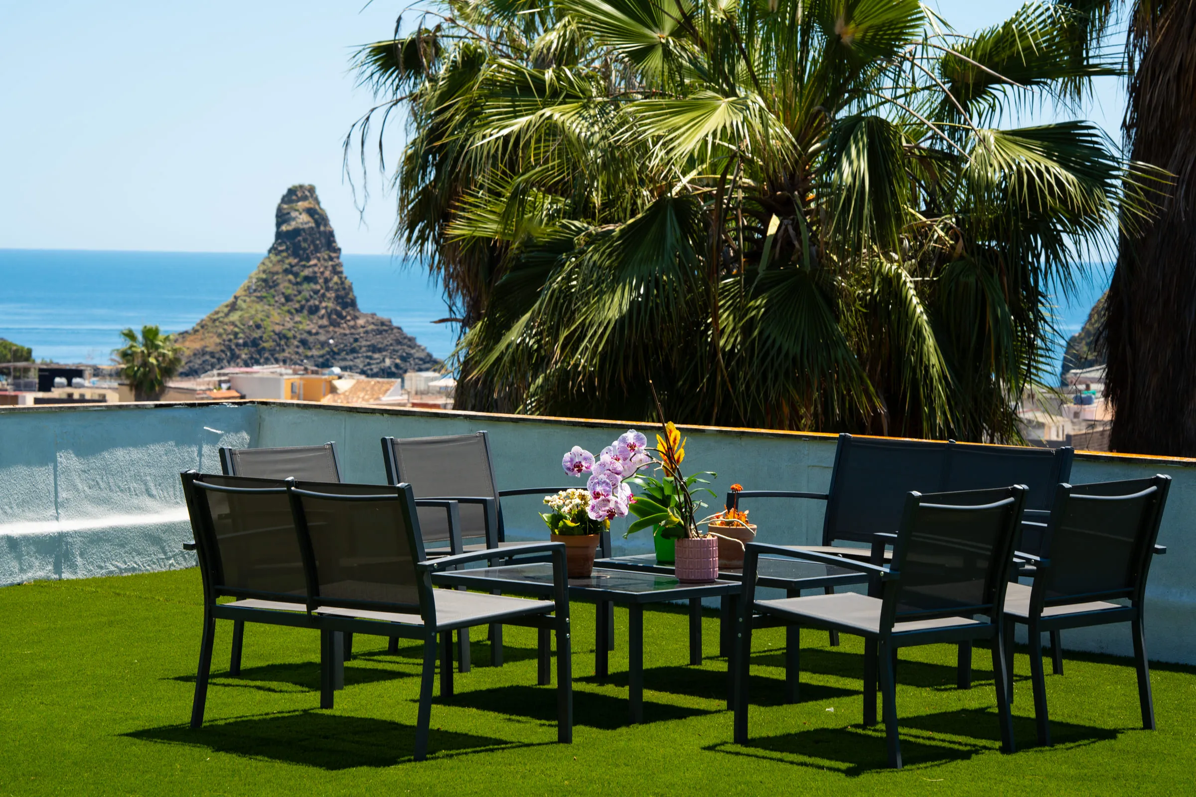 The apartment's terrace with table and armchairs, the faraglione rising from the sea beyond the palms.
